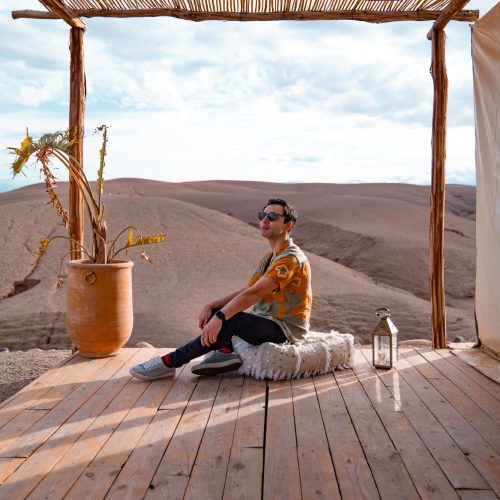 Man with sunglasses sitting on a wooden deck in a sunny outdoor setting. Agafay desert, Morocco.