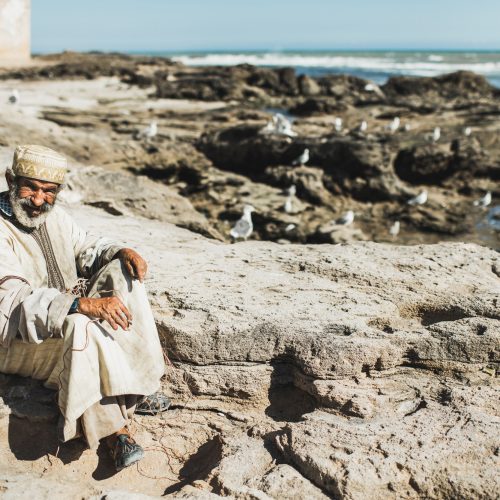 Old arab fisherman in traditional white cape and skullcap smoking and rest on coast of Essaouira